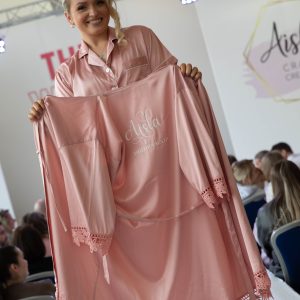 Blond haired woman on a catwalk holding a blush pink lace edge robe personalised in white text