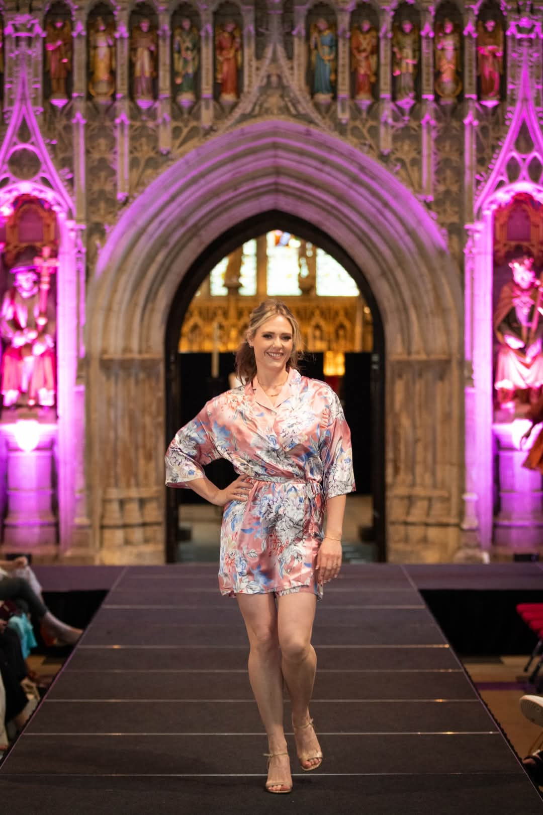 Blond haired woman wearing a pink floral satin silk robe, on a catwalk in a cathedral.